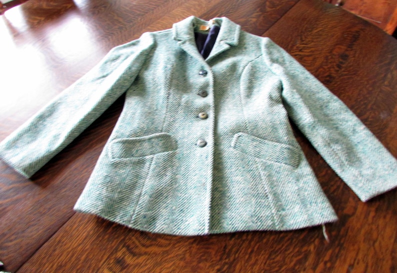 May include: A long-sleeved blazer in a light teal and white herringbone pattern. The jacket features a notched lapel, four dark buttons down the center front, and two horizontal welt pockets at the waist. The interior is lined with dark fabric.