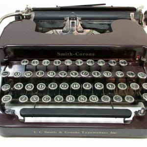 May include: A vintage Smith-Corona typewriter in a deep burgundy color. The keyboard features round, silver keys with black lettering. The text "Smith-Corona" is visible above the keys, and "L. C Smith & Corona Typewriters Inc" is below.