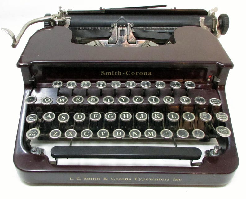 May include: A vintage Smith-Corona typewriter in a deep burgundy color. The keyboard features round, silver keys with black lettering. The text "Smith-Corona" is visible above the keys, and "L. C Smith & Corona Typewriters Inc" is below.