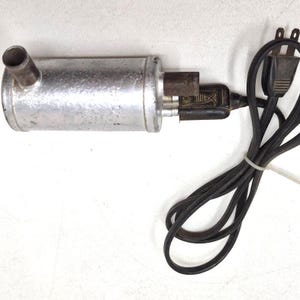May include: A vintage silver-colored metal appliance with a cylindrical body and a short, curved spout. It has a black power cord with a two-prong plug. The cord is bundled with a white tie.