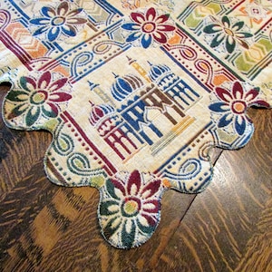 May include: A decorative, beige table runner with a scalloped edge. The runner features a repeating floral and geometric pattern in shades of blue, red, green, and orange. A central design depicts a building with domes.
