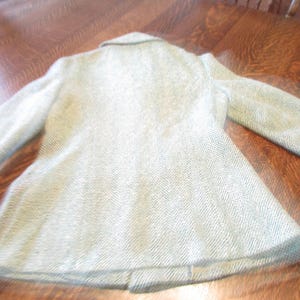 May include: A light-colored blazer featuring a subtle herringbone pattern throughout the fabric. The garment includes long sleeves, a structured collar, and a tailored fit that tapers slightly at the waist, showcasing a classic design.