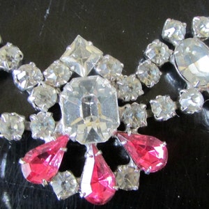 May include: A silver necklace with clear and pink rhinestones. The necklace features a central cluster of larger rhinestones with smaller rhinestones surrounding it. The necklace is adorned with teardrop-shaped pink rhinestones.