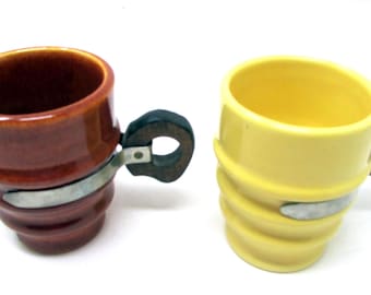 May include: Two ceramic mugs, one brown and one yellow, with a distinctive design. Each mug features a ridged body, a wooden handle, and a metal band. The brown mug has a dark interior, while the yellow mug has a light interior.