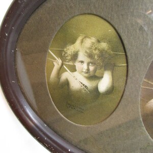 K&ouml;nnte beinhalten: Ein ovaler Bilderrahmen im Vintage-Stil mit einem braunen Holzrahmen und einer Glasscheibe. Der Rahmen enth&auml;lt drei ovale Bilder. Das mittlere Bild ist ein sepiafarbenes Foto eines Cherubins, der einen Pfeil h&auml;lt. Der Text "Copyright 1902 by A.T.C. London" ist unter dem Bild gedruckt.