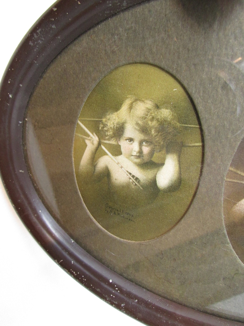 K&ouml;nnte beinhalten: Ein ovaler Bilderrahmen im Vintage-Stil mit einem braunen Holzrahmen und einer Glasscheibe. Der Rahmen enth&auml;lt drei ovale Bilder. Das mittlere Bild ist ein sepiafarbenes Foto eines Cherubins, der einen Pfeil h&auml;lt. Der Text "Copyright 1902 by A.T.C. London" ist unter dem Bild gedruckt.