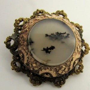 May include: A vintage brooch with a round, white agate stone set in a gold-toned filigree frame. The stone has black dendritic inclusions.