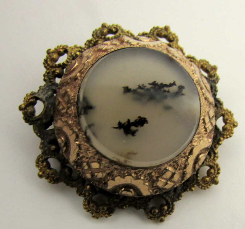 May include: A vintage brooch with a round, white agate stone set in a gold-toned filigree frame. The stone has black dendritic inclusions.