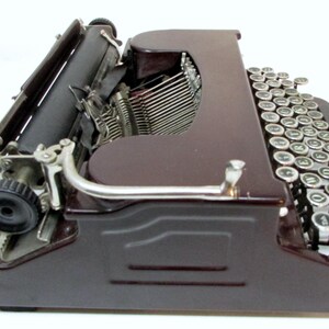 May include: A vintage brown typewriter with a black roller and silver keys. The keys are round with white lettering. The typewriter has a classic design, with a curved handle and a metal frame.