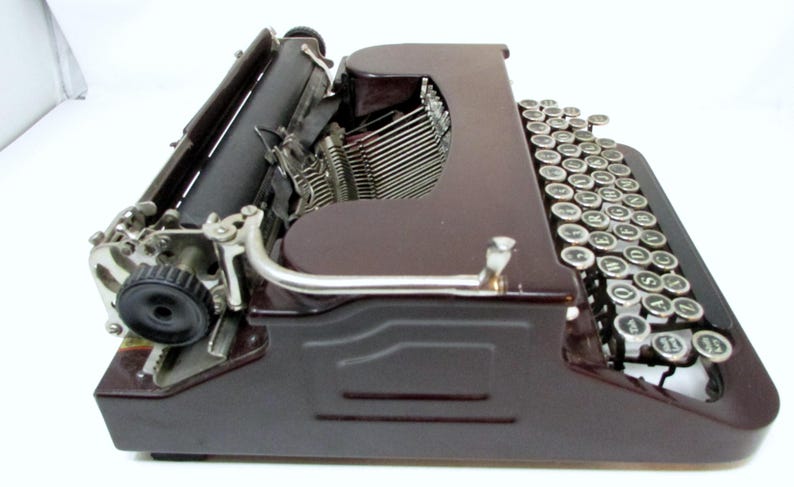 May include: A vintage brown typewriter with a black roller and silver keys. The keys are round with white lettering. The typewriter has a classic design, with a curved handle and a metal frame.