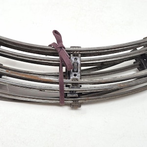 May include: A stack of curved model train tracks made of metal. The tracks are held together with a maroon ribbon. The tracks are a dark gray color and appear to be vintage.