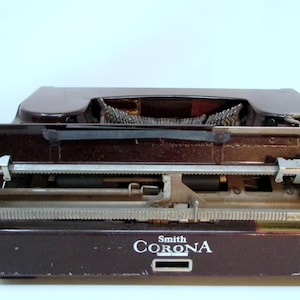 May include: A vintage Smith Corona typewriter in a dark brown finish. The typewriter features a classic design with a carriage return lever and a keyboard. The "Smith Corona" logo is visible on the front of the machine.