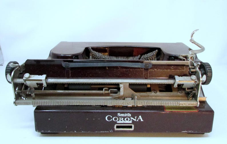 May include: A vintage Smith Corona typewriter in a dark brown finish. The typewriter features a classic design with a carriage return lever and a keyboard. The "Smith Corona" logo is visible on the front of the machine.