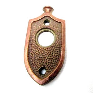 May include: A vintage copper-toned door plate with a shield shape. The plate has a textured surface and three holes: a large central hole and two smaller ones. The top has a small, rounded protrusion.