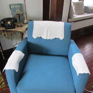 May include: A teal armchair with white lace doilies on the back and armrests. The doilies have a scalloped edge and embroidered floral details. The chair is in a room with a vintage telephone and a book titled "Art Deco".