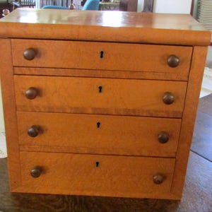 Könnte beinhalten: Eine kleine Holzkommode mit vier Schubladen. Die Kommode hat eine warme, hellbraune Farbe mit glänzendem Finish. Jede Schublade hat einen kleinen, runden, dunkelbraunen Knopf und ein Schlüsselloch. Die Kommode steht auf einer Holzoberfläche.