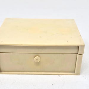 May include: A small, rectangular, off-white box with a single drawer. The box has a simple design with a small, round knob on the drawer. The box is made of a solid material and appears to be vintage.