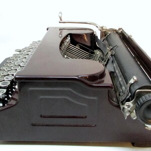 May include: Side view of a vintage maroon typewriter with silver keys and a black roller. The typewriter has a classic design with a curved body and a lever on top. The keys are arranged in a standard QWERTY layout.