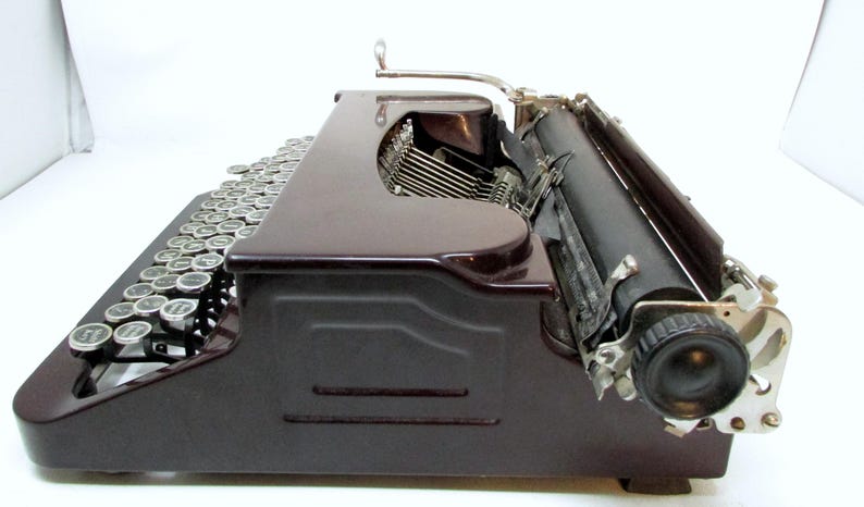 May include: Side view of a vintage maroon typewriter with silver keys and a black roller. The typewriter has a classic design with a curved body and a lever on top. The keys are arranged in a standard QWERTY layout.
