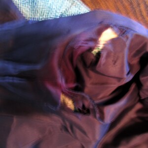 May include: A dark purple garment lining featuring a visible tear along the seam, revealing the lighter colored fabric underneath. The smooth, dark purple material is bunched and folded, showing the damaged area where the stitching has separated.