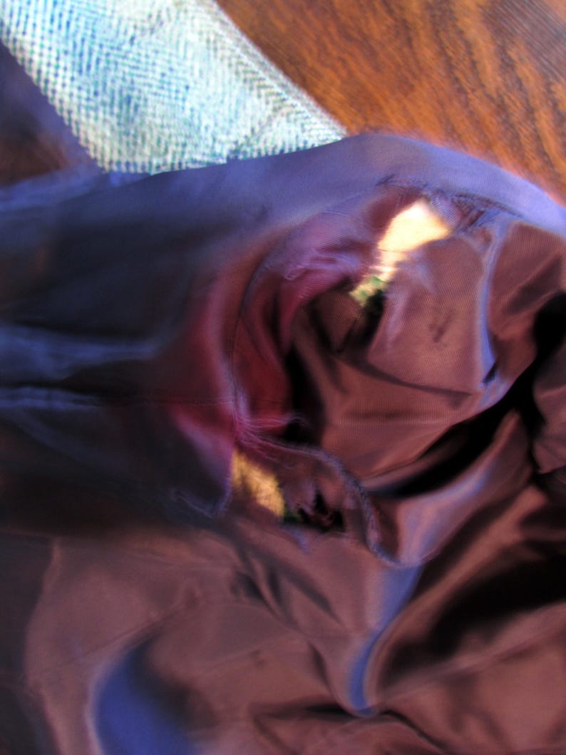 May include: A dark purple garment lining featuring a visible tear along the seam, revealing the lighter colored fabric underneath. The smooth, dark purple material is bunched and folded, showing the damaged area where the stitching has separated.