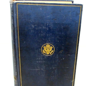 Op de afbeelding: Een marineblauw hardcover boek met gouden rand en een gouden embleem. De kaft van het boek heeft een licht versleten uiterlijk. Het embleem is een goudkleurige afbeelding van het Grote Zegel van de Verenigde Staten.