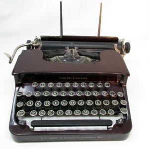 May include: A vintage Smith-Corona typewriter in a deep burgundy color. The keyboard features round, silver keys with black lettering. The text "Smith-Corona" is visible above the keys, and "L. C. Smith & Corona Typewriters Inc" is below.