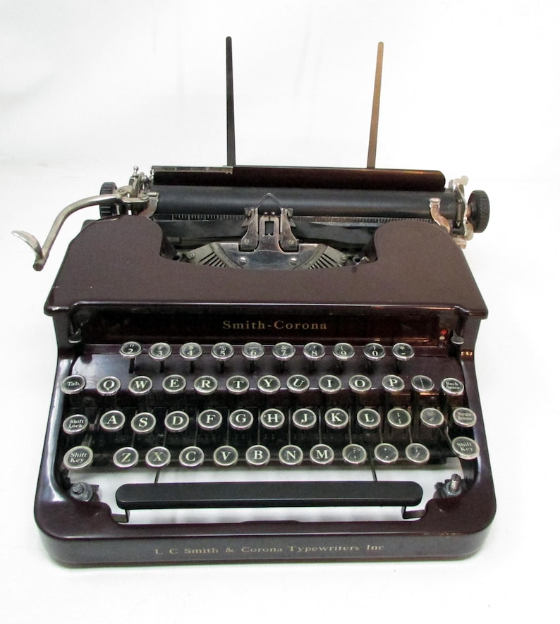 May include: A vintage Smith-Corona typewriter in a deep burgundy color. The keyboard features round, silver keys with black lettering. The text "Smith-Corona" is visible above the keys, and "L. C. Smith & Corona Typewriters Inc" is below.