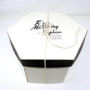 May include: A hexagonal white hat box with a black lid edge and a white string tied around the center. The lid features black printed text that reads Rose Millinery at Johnson, with the locations Tacoma, Lakewood, and Yakima listed below.