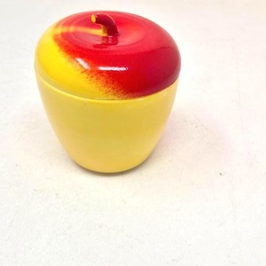 Könnte beinhalten: Ein gelber, apfelförmiger Behälter mit einem rot-gelben Deckel. Der Deckel hat einen kleinen Stiel. Der Behälter ist wahrscheinlich aus Keramik oder Kunststoff und soll einem realistischen Apfel ähneln.