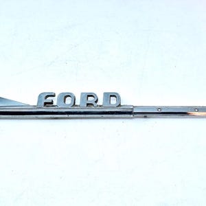 Puede incluir: Adorno de capó "FORD" cromado vintage con un diseño aerodinámico. El adorno tiene un extremo puntiagudo y una aleta triangular. La palabra "FORD" está en letras de bloque.
