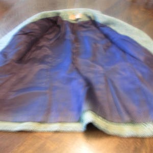 May include: A light green textured coat with a dark blue interior lining. The garment features a collar and long sleeves, with the front open to reveal the smooth, dark blue fabric inside. The outer material has a visible woven pattern.