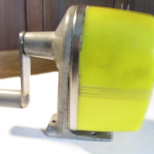 May include: A manual pencil sharpener featuring a bright yellow plastic casing with horizontal grooved lines, a metallic silver-toned frame, and a grey plastic crank handle. The device has a sturdy base with visible screw holes for mounting.