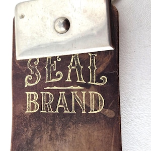Puede incluir: Un cuero para afilar vintage con las palabras "SEAL BRAND" en letras doradas. El cuero tiene una pieza final de metal y un cierre plateado.