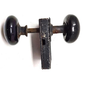 以下が含まれることがあります： 2つの黒い楕円形のドアノブが金属棒と長方形のラッチ機構で接続された、アンティークのドアハードウェアセット。 金具には経年劣化と摩耗の兆候が見られ、ノブは光沢のある仕上がりです。