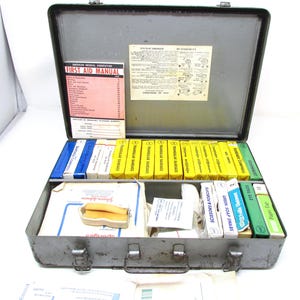 May include: Open metal first aid kit containing medical supplies. The kit includes bandages, ointments, scissors, and a first aid manual. The kit is grey with a handle and latches.