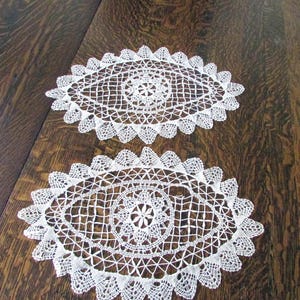 May include: Two white, oval-shaped doilies with intricate lace patterns. The doilies have a central floral design and a scalloped edge. They are placed on a dark brown wooden surface.