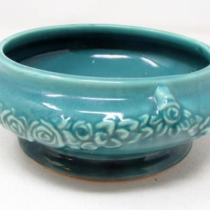 May include: A round, teal-colored ceramic bowl featuring a raised, embossed floral pattern around the exterior. The bowl has a smooth, glossy finish, a wide opening, and a sturdy base, showcasing a consistent monochromatic blue-green glaze.