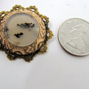 May include: A gold-toned metal brooch with a round, white agate stone set in the center. The stone has black dendritic patterns. The brooch is surrounded by a decorative, filigree design.