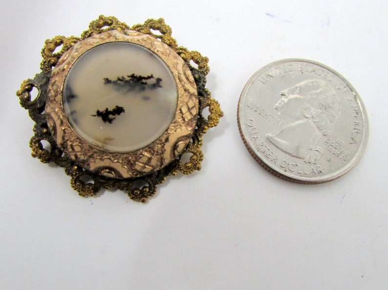 May include: A gold-toned metal brooch with a round, white agate stone set in the center. The stone has black dendritic patterns. The brooch is surrounded by a decorative, filigree design.