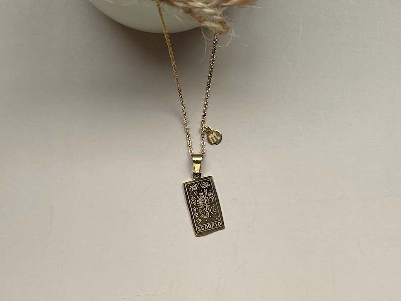 May include: Gold necklace with a pendant featuring a zodiac sign, Scorpio, and a small charm with the letter 'm'.