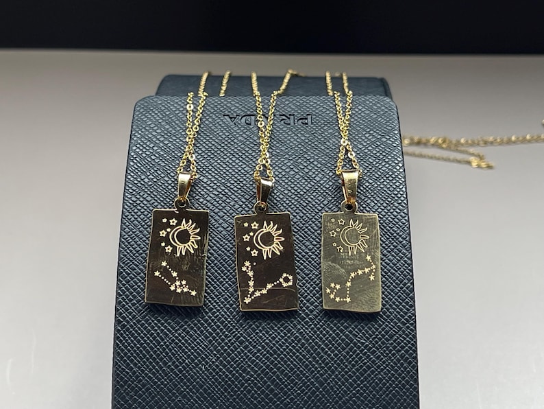 May include: Three gold necklaces with rectangular pendants featuring a crescent moon, sun, and stars design. Each pendant has a constellation design.