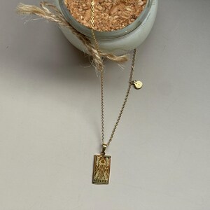 May include: A gold chain necklace with a pendant featuring a tarot card design. The pendant is rectangular and has a detailed image of a figure with wings. The chain has a small round charm attached.