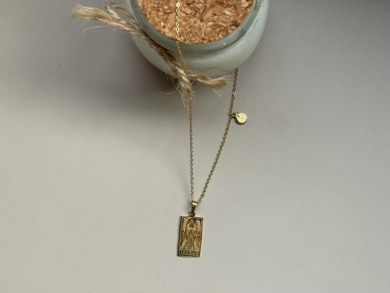May include: A gold chain necklace with a pendant featuring a tarot card design. The pendant is rectangular and has a detailed image of a figure with wings. The chain has a small round charm attached.