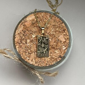 May include: Gold necklace with a rectangular pendant featuring a black and white illustration of two fish and the word "Pisces".