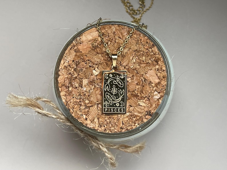 May include: Gold necklace with a rectangular pendant featuring a black and white illustration of two fish and the word "Pisces".