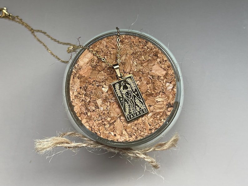 May include: Gold necklace with a pendant featuring a black and white illustration of a tarot card with the word "Taurus" on it.
