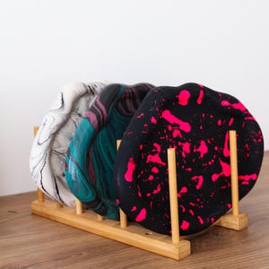 Könnte beinhalten: Drei dekorative Tabletts mit einzigartigen Marmordesigns in Weiß, Türkis und Schwarz mit pinkfarbenen Spritzern. Die Tabletts werden in einem Naturholzregal ausgestellt, das ihre künstlerischen Muster und Farben zeigt. Ein stilvolles Wohnaccessoire.