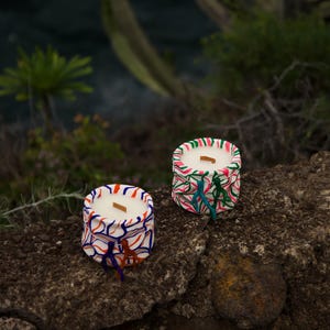 Pode incluir: Duas velas pequenas e redondas com cera branca e desenhos abstratos coloridos. As velas estão sobre uma superfície rochosa.