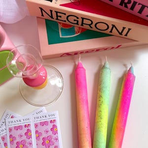 Peut inclure: Une collection de bougies colorées et d'articles sur le thème des cocktails. Les bougies sont vibrantes, avec un dégradé de rose, de jaune et de vert. Des boîtes de cocktails avec les mots "NEGRONI" et "APEROL SPRITZ" sont également présentes.